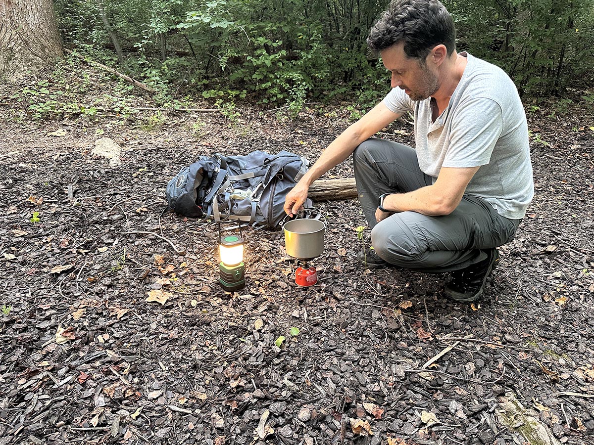 A person using a camping stove in the woods next to the best LED lantern option