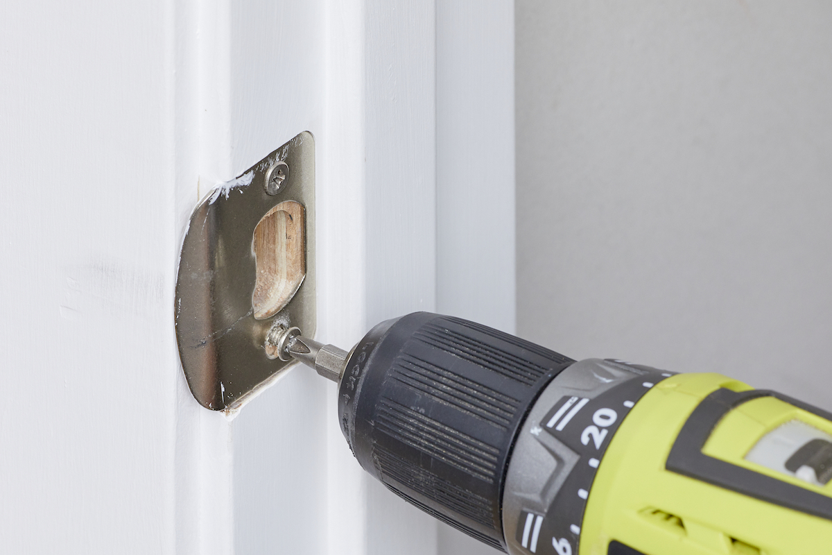 Woman uses an electric screwdriver to remove screws from strike plate in a door frame.