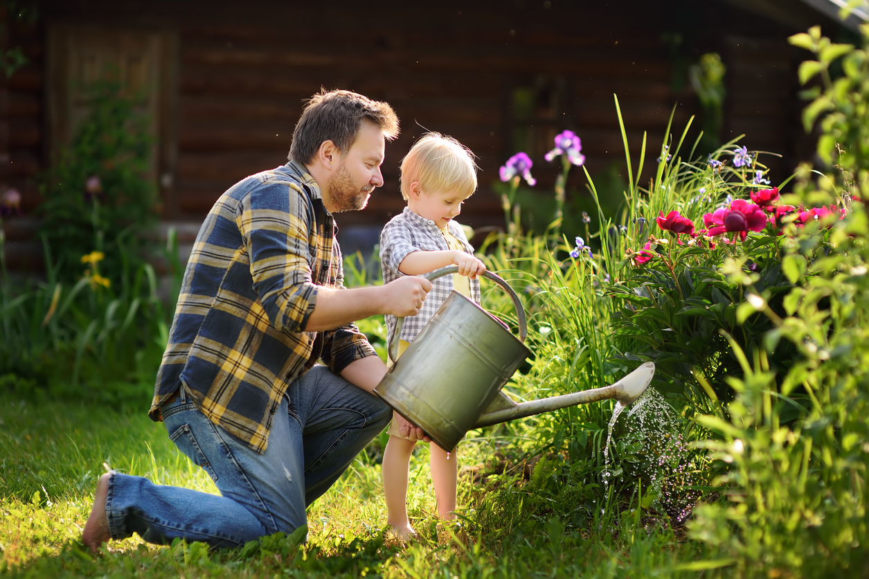 Middle age man and his little son watering flowers in the garden at summer sunny day. Gardening activity with little kid and family