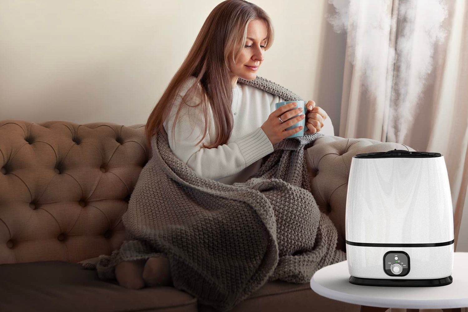 A person on the couch wrapped in a blanket and holding a mug with the best humidifier for large rooms sitting on a coffee table nearby