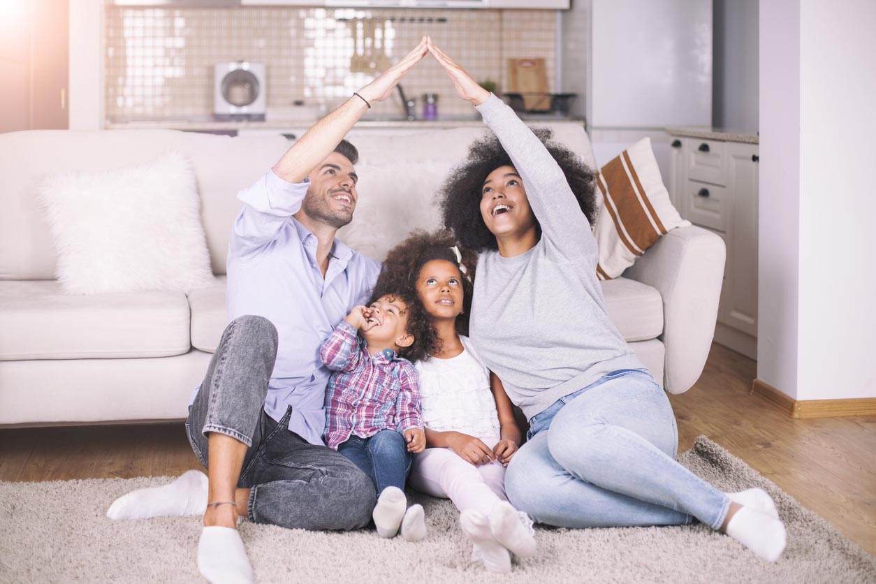 A man and a woman sit on a living room floor with two children and form a roof with their arms over them. 