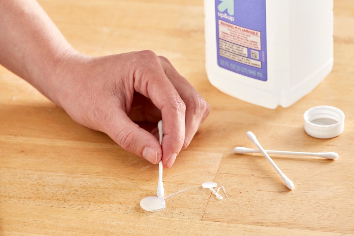 Woman uses a Q-tip to apply alcohol to a hot glue spot on a wood table.