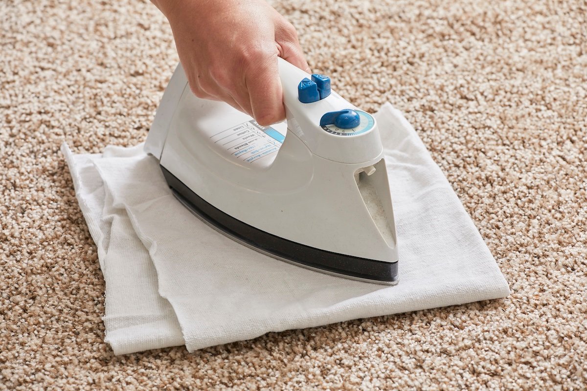 Woman places an iron atop a piece of folded white cloth that's on top of a light brown carpet.