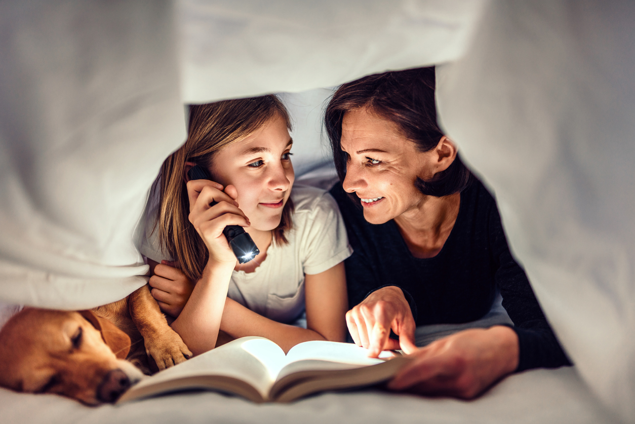 Mother, daughter and dog laying on the bed under blanket holding flashlight and reading book late at night.