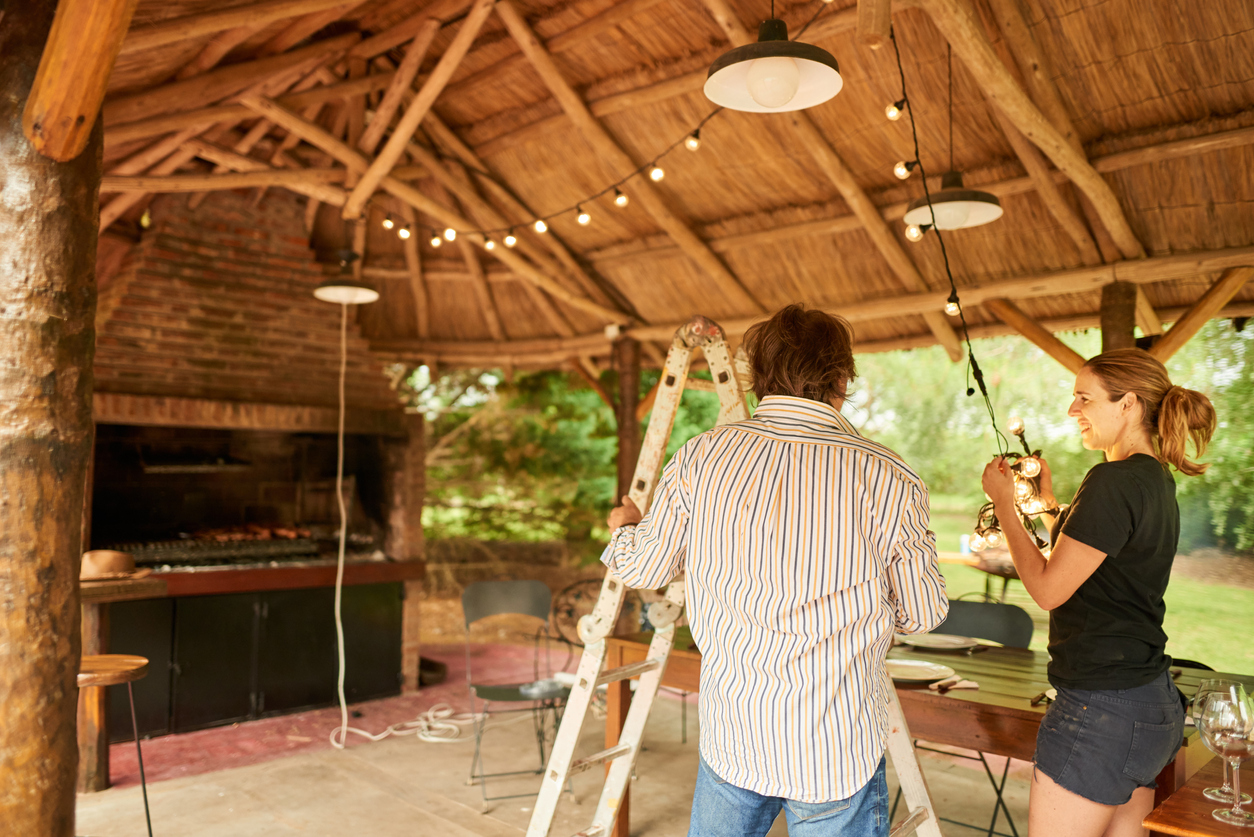 Man and woman using ladder to hang string lights under covered patio