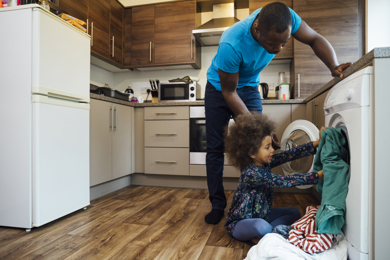 Wide shot of a mixed race girl sitting on the floor in the kitchen and helping her father put the washing into the washing machine.