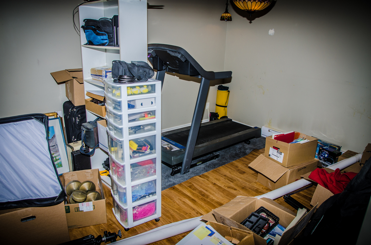 A cluttered room in a finish basement that contains gym equipment, and cardboard boxes and plastic organizers filled with personal belongings.