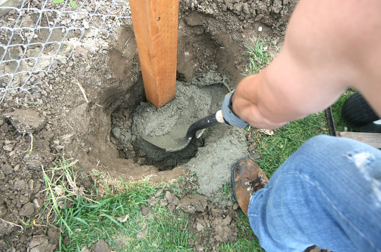 Person digging hole around concrete fence post base.