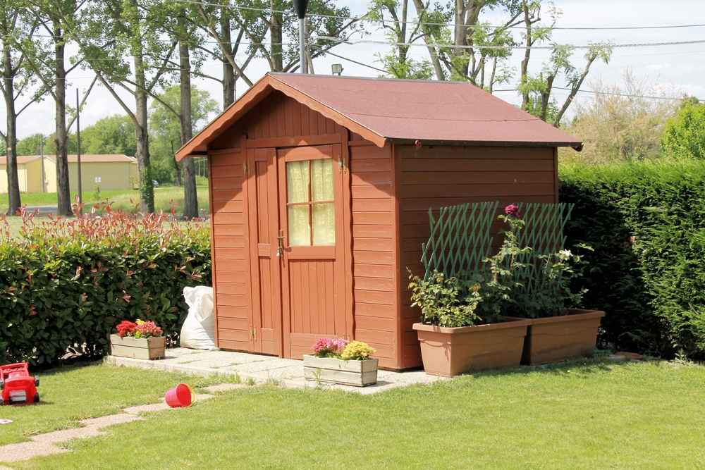 A finished shed on a patio with hedges in the background and kid's toys in the foreground.