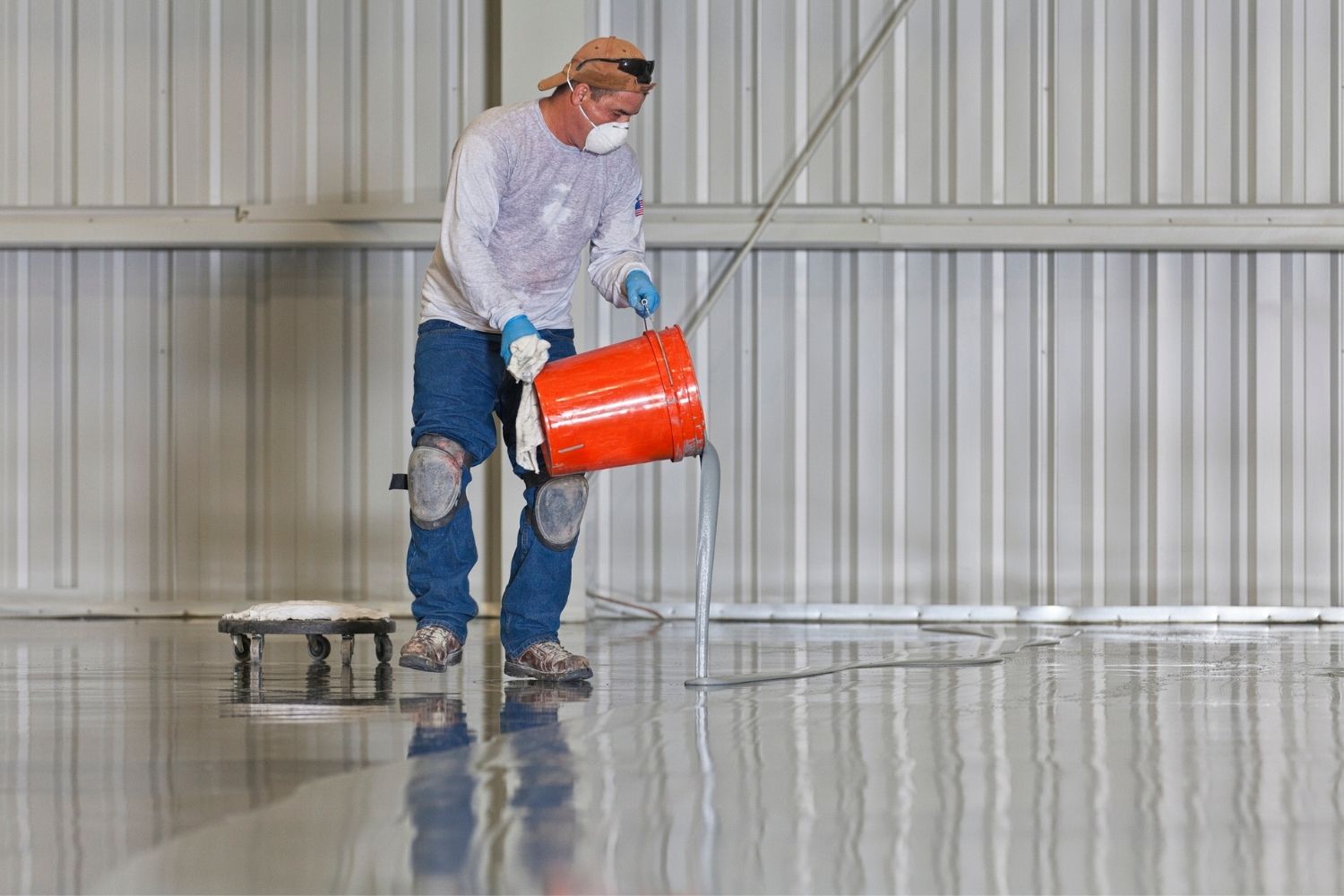 A worker wearing a dust mask pours epoxy coating from a red bucket onto a garage floor.