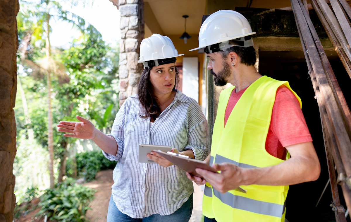 A homeowner reviews a building permit with a contractor on an iPad while both wear hardhats.