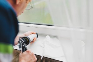 Backview of an elderly man using a silicone tube of caulk on a windowsill.
