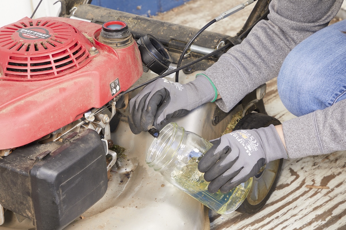Woman wearing work gloves empties gas from a lawn mower into a glass jar.