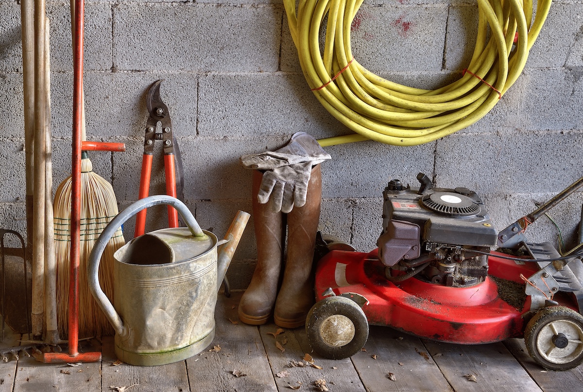 Lawn mower, watering can, extension cord, and other lawn tools in storage against cement wall.