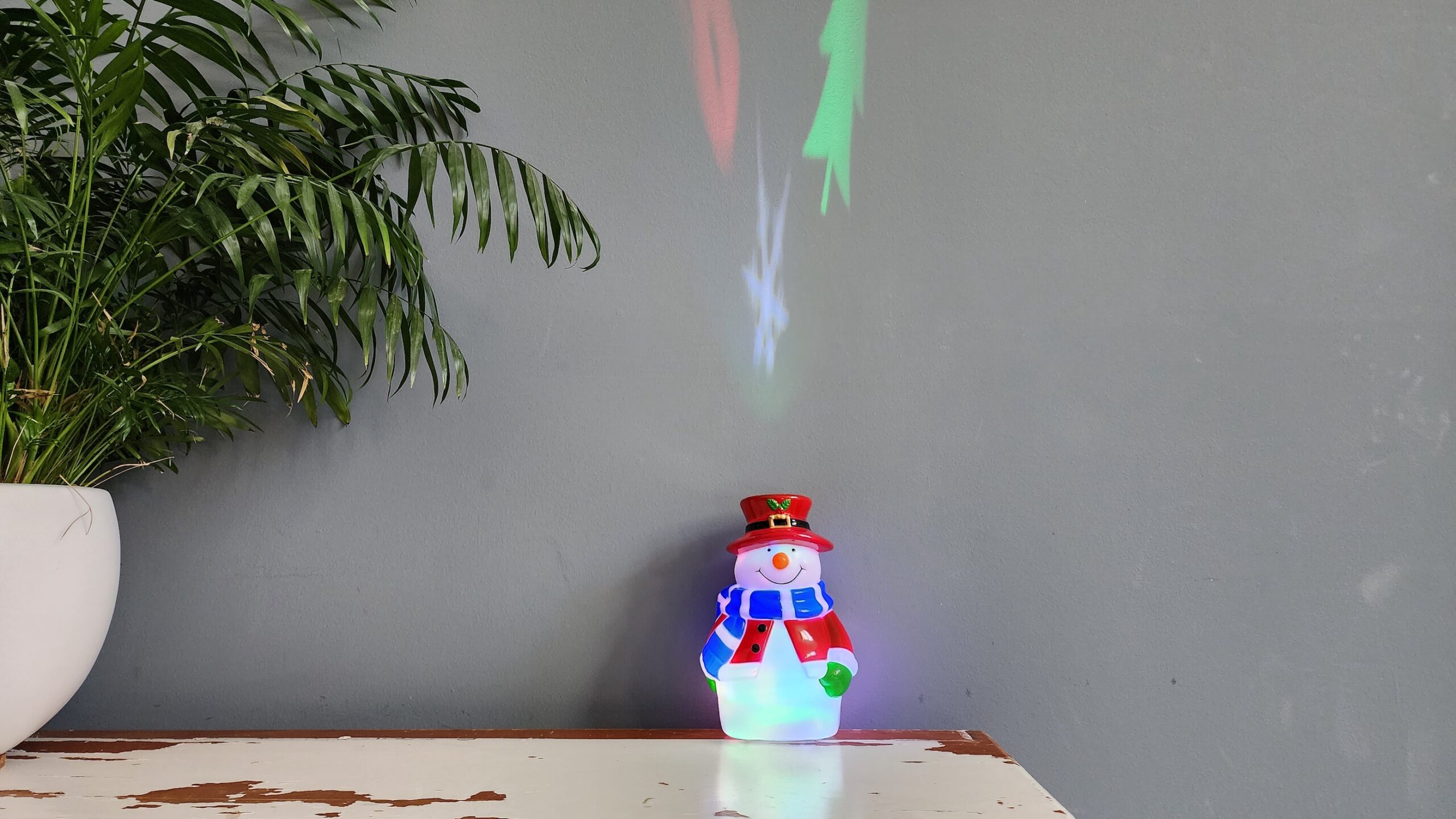 The Yocuby Snowman Christmas Light Projector on an antique table and projecting a red, blue, and green holiday scene onto the wall above.