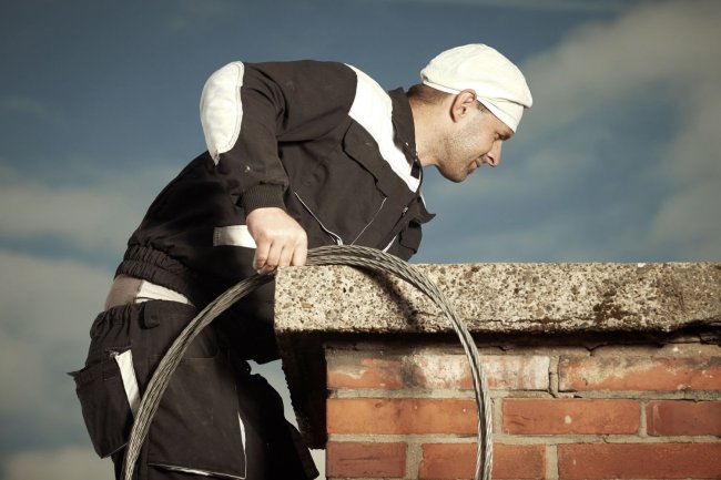 A man dressed in black reaches down into a brick chimney on top of a roof.