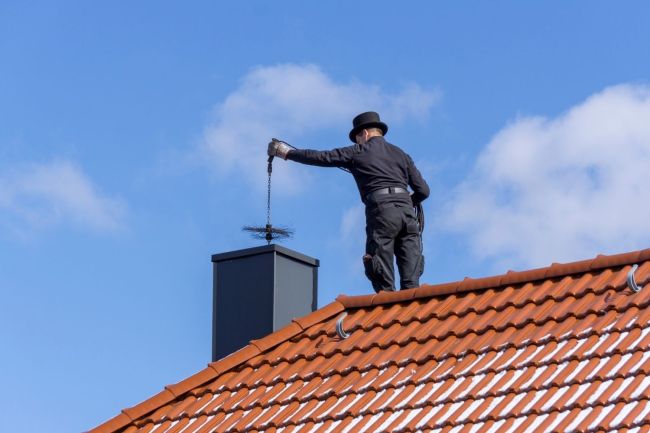 A worker dressed in black stands on a red tile roof and prepares to clean a chimney.