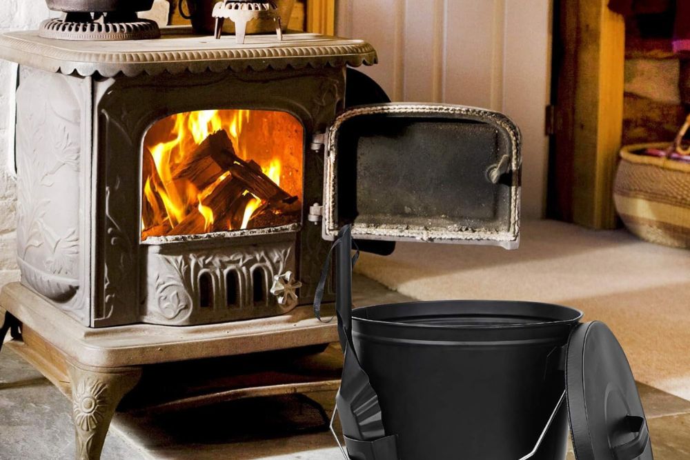 The F2C Ash Bucket With Lid and Shovel on the floor next to a wood-burning stove with its door open to show a fire burning inside.