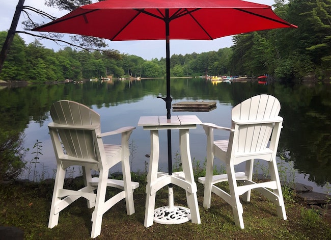 Two tall white Adirondack chairs with small table and red patio umbrella in front of lake