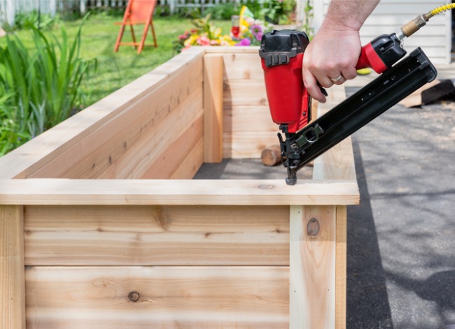 Person uses a red finish nailer on a new wooden raised garden bed.