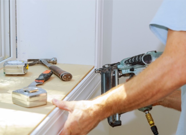 A man uses a finish nailer to nail in trim around a window with an assortment of tools nearby.
