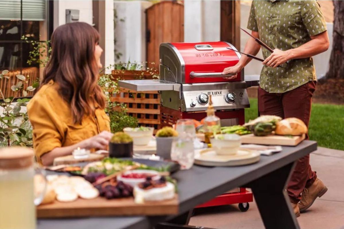 A woman sitting at an outdoor table full of food and drinks while a man stands holding tongs next to the best gas grill under $500