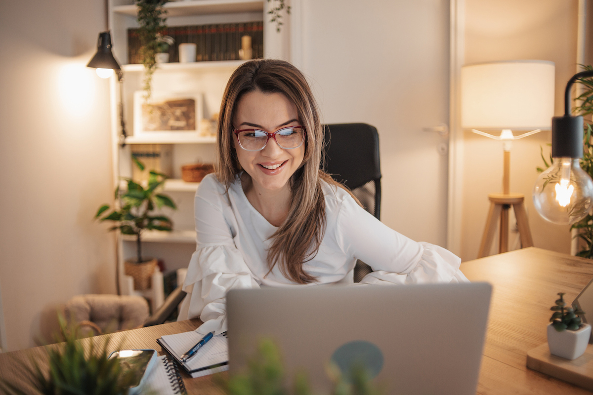 Woman with glasses working on laptop in home office with lamps next to her and behind her.