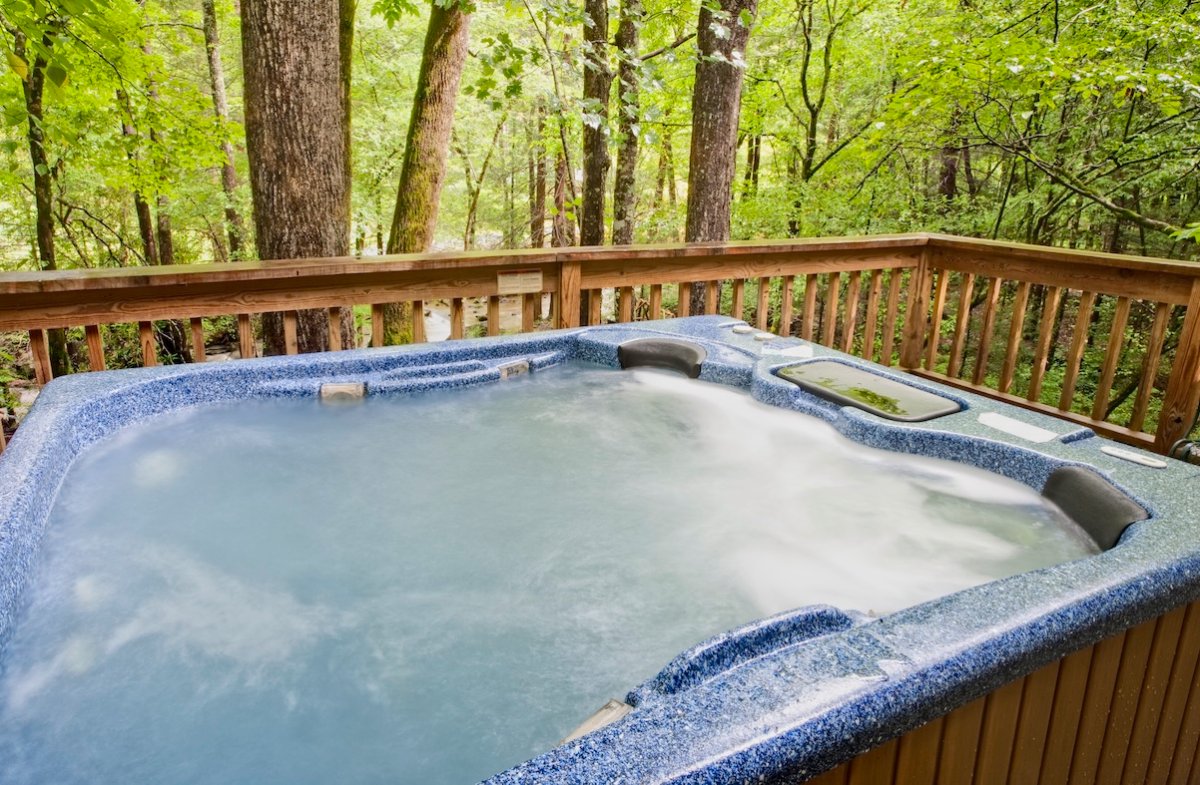 Full hot tub on the deck of a wood cabin, with verdant trees in the background.