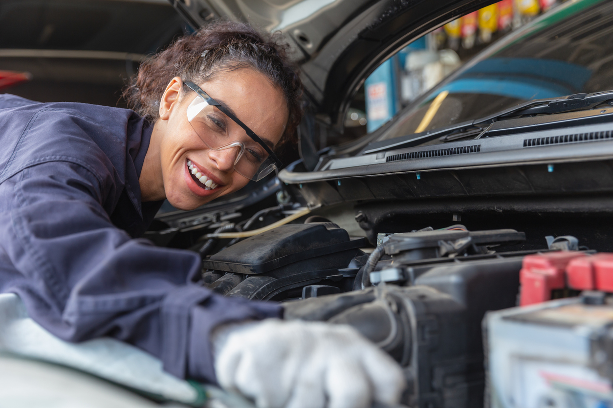 woman mechanic wearing gloves and goggles reaching for car battery engine hood open