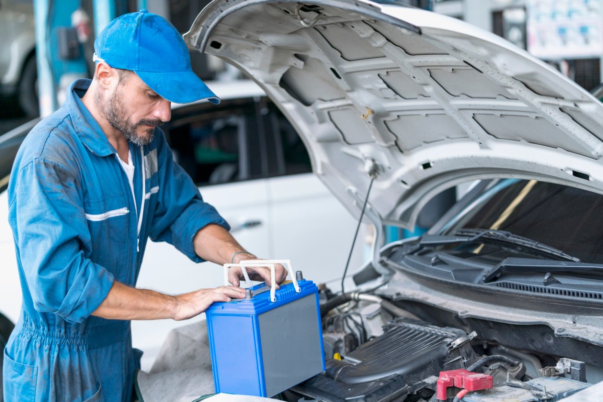 how to change a cary battery mechanic in blue jumpsuit holding car battery with engine hood open