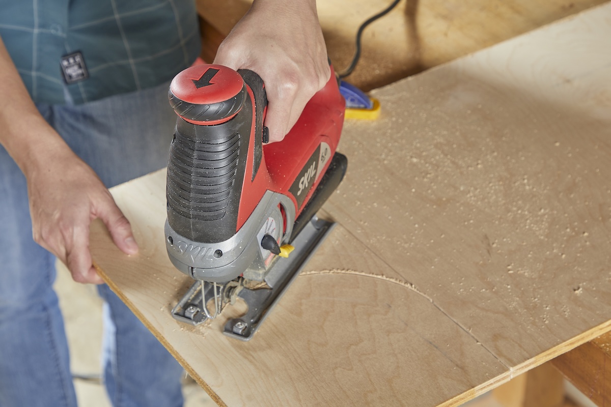 Woman uses a jigsaw to cut a curve into a piece of plywood.