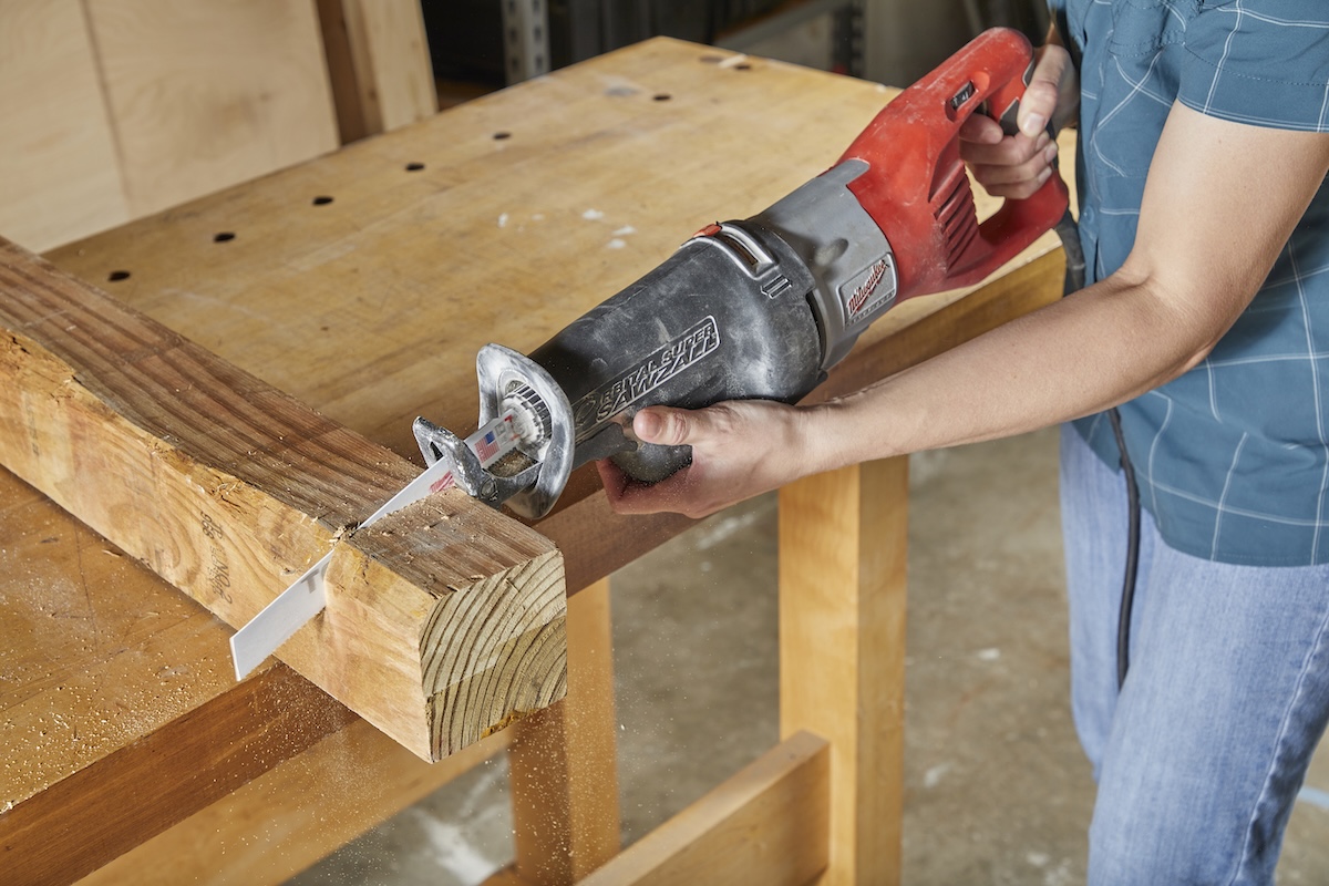 Woman uses a reciprocating saw to cut through a thick wood post. 