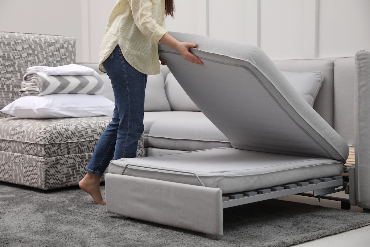 Woman making sleeping place for guest on a grey chaise sofa bed in living room.