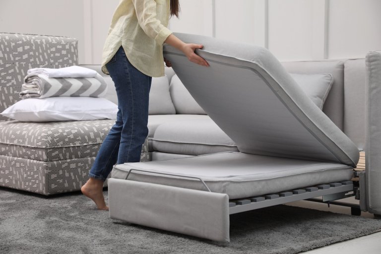 Woman making sleeping place for guest on a grey chaise sofa bed in living room.
