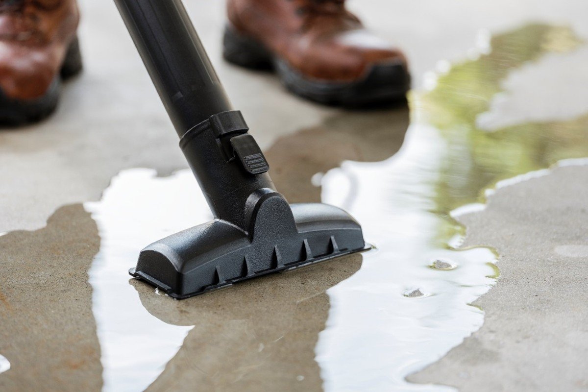 A person is using a wide, T-shaped attachment with their shop vac to remove water from the flat floor.