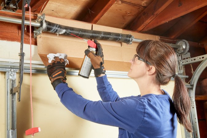 Woman standing on step stool and lubricating garage door spring with silicone spray.