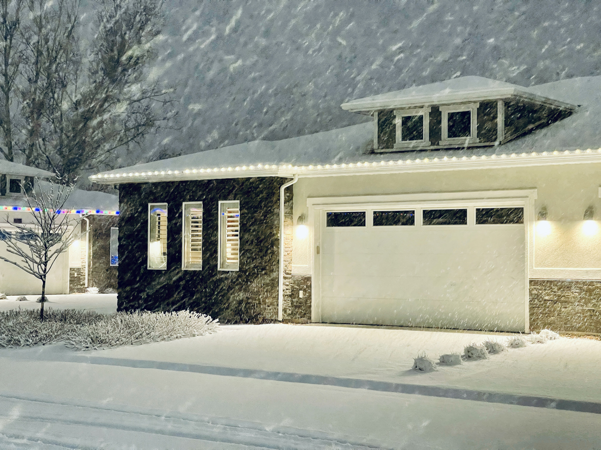Midcentury style house in a snowstorm at dusk, with holiday lights on next door house.