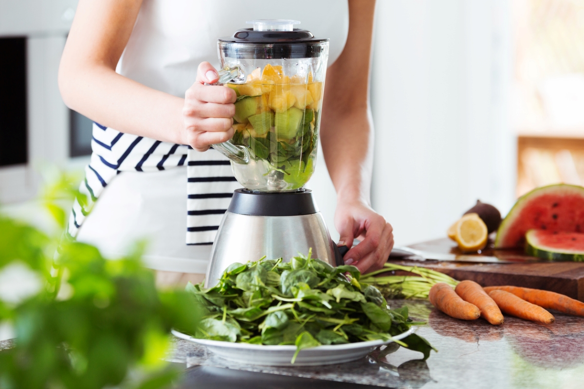 Woman wearing striped skirt using kitchen blender with vegetables all over table.