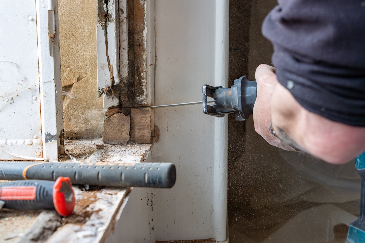Man uses a reciprocating saw to cut through wood and nails around a window frame.
