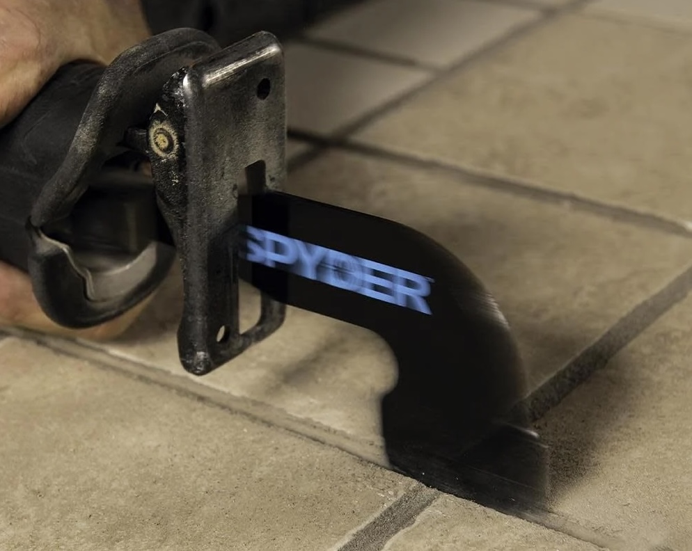 A reciprocating saw with a Spyder grout removal attachment saws grout from between beige tiles.