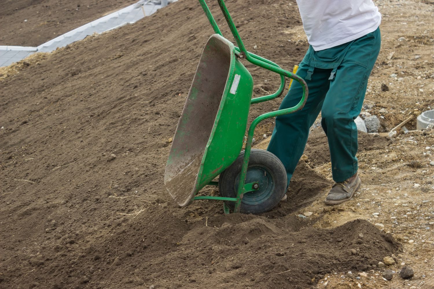 A worker empties out a wheelbarrow with fill dirt onto a sloped area.