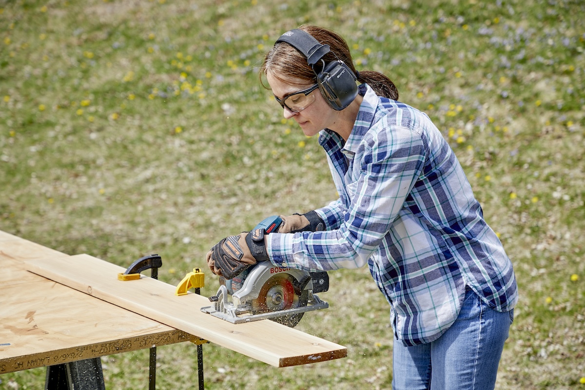 Woman wearing ear and eye protection cuts a 2 x 4 with a circular saw at an outdoor workbench.