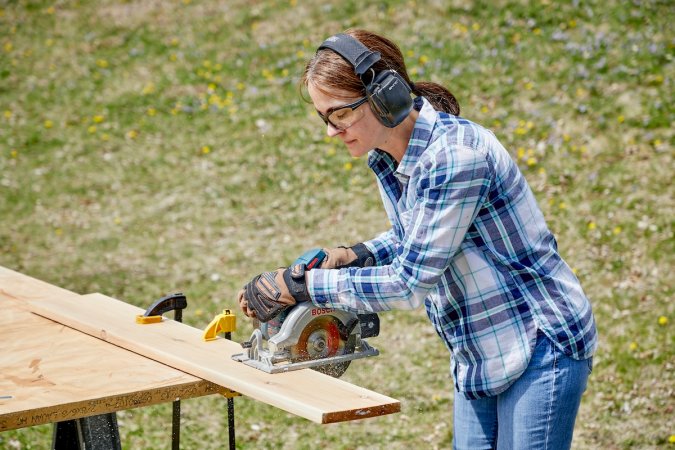Woman wearing ear and eye protection cuts a 2 x 4 with a circular saw at an outdoor workbench.