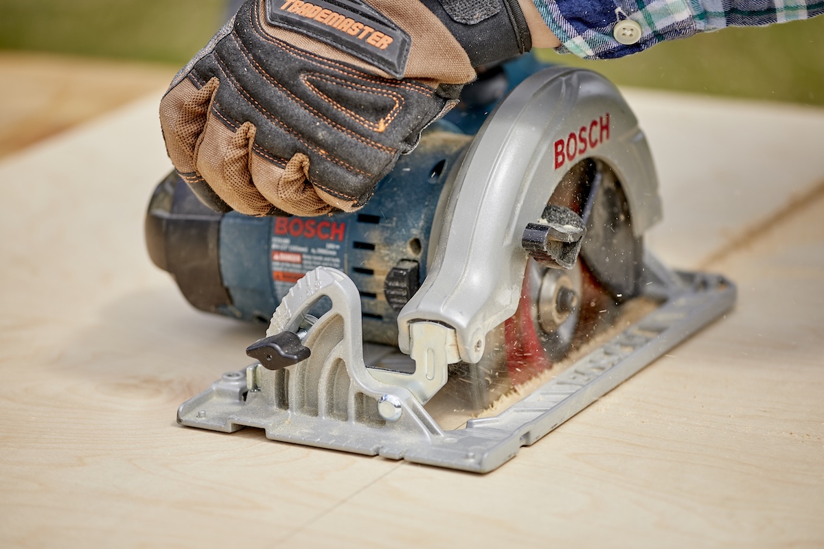 Close up of person wearing work gloves usaing a circular saw to cut through plywood.