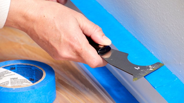 Woman uses a painter's tool to press painter's tape over a baseboard to protect it before painting.