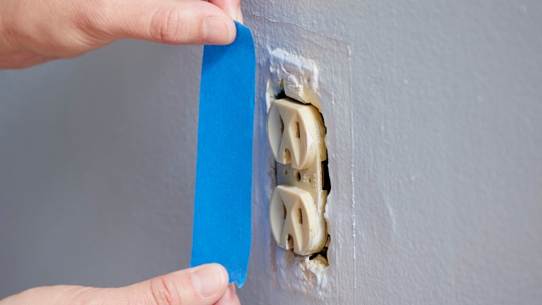 Woman uses blue painter's tape to cover electrical outlet before painting a wall.