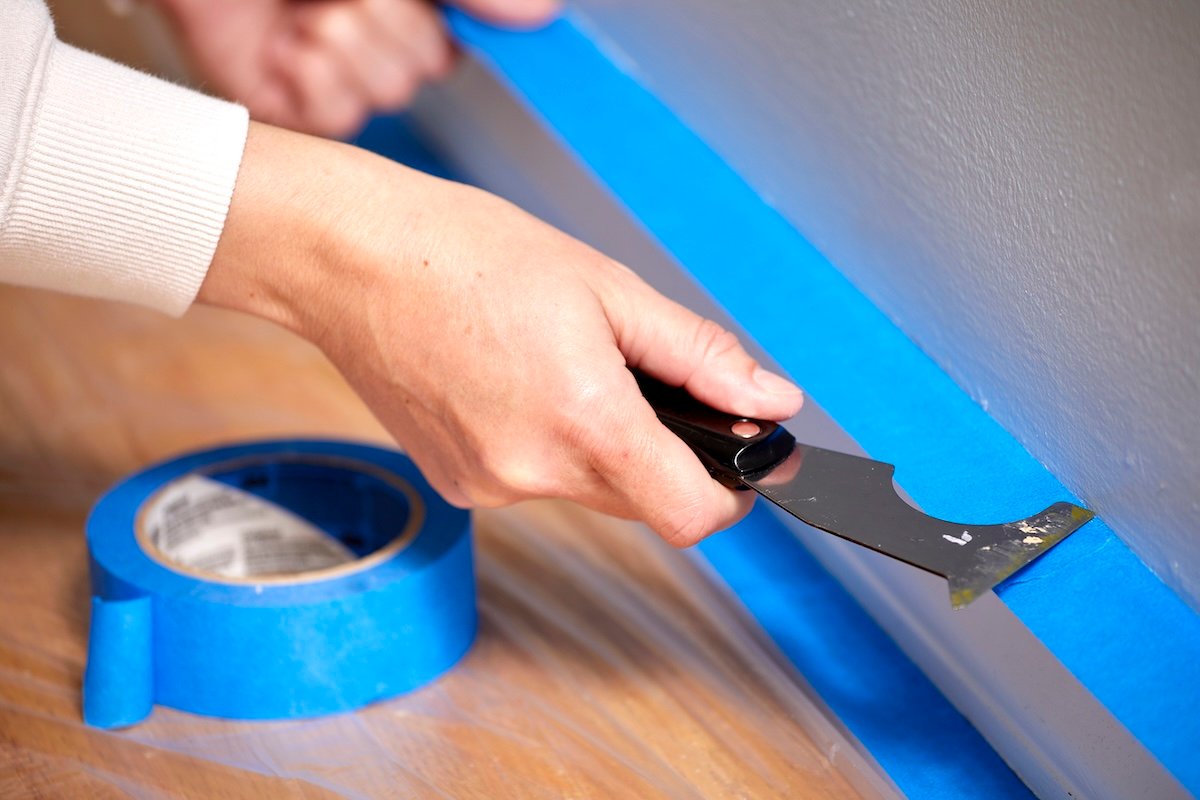 Woman uses a painter's tool to smooth painter's tape onto a baseboard.