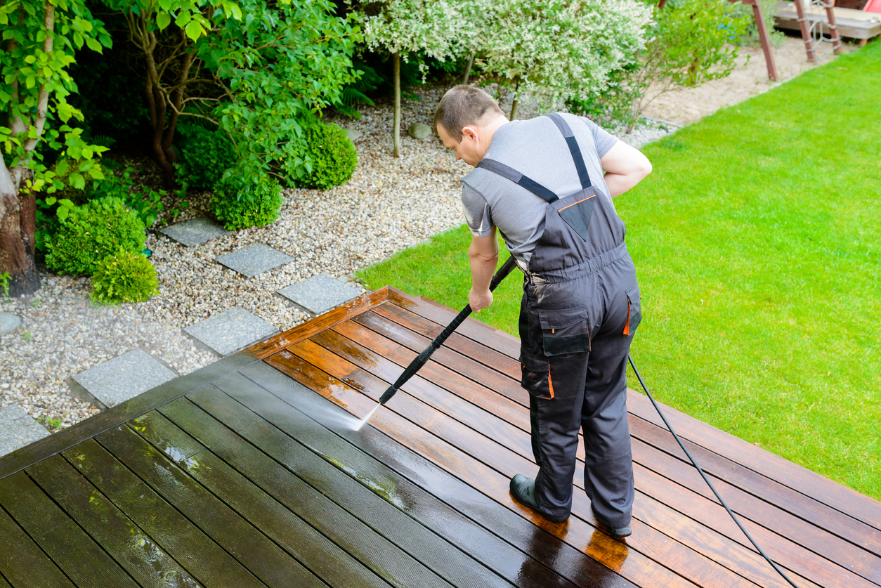 Man cleans a low wood deck in a backyard with a power washer.