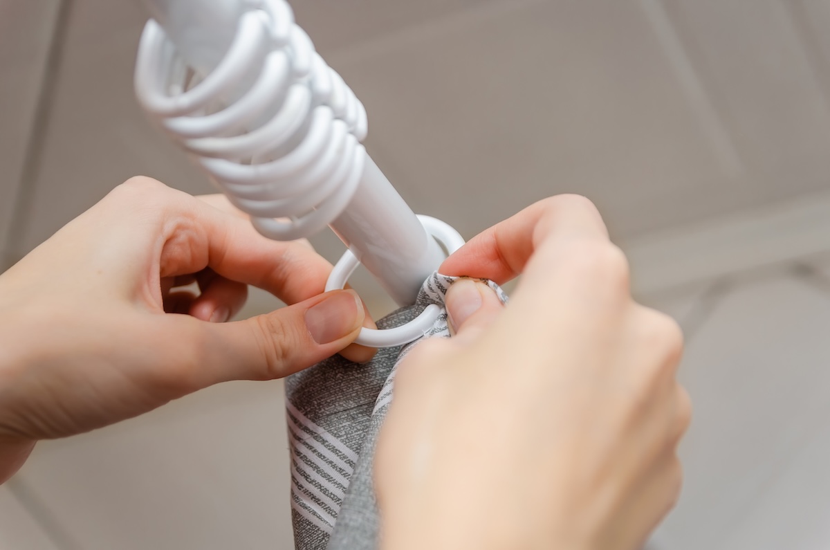 Closeup of women's hands hanging a gray shower curtain on white shower curtain rings.