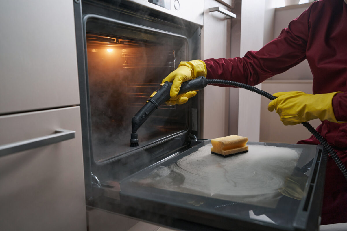 A person with yellow rubber gloves uses a steam cleaner to clean the interior of their oven. 